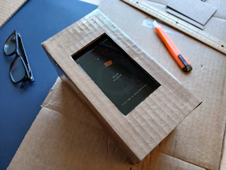 A close-up of a constructed cardboard box with a hole cut in the center for a mobile phone, the meditation timer being used